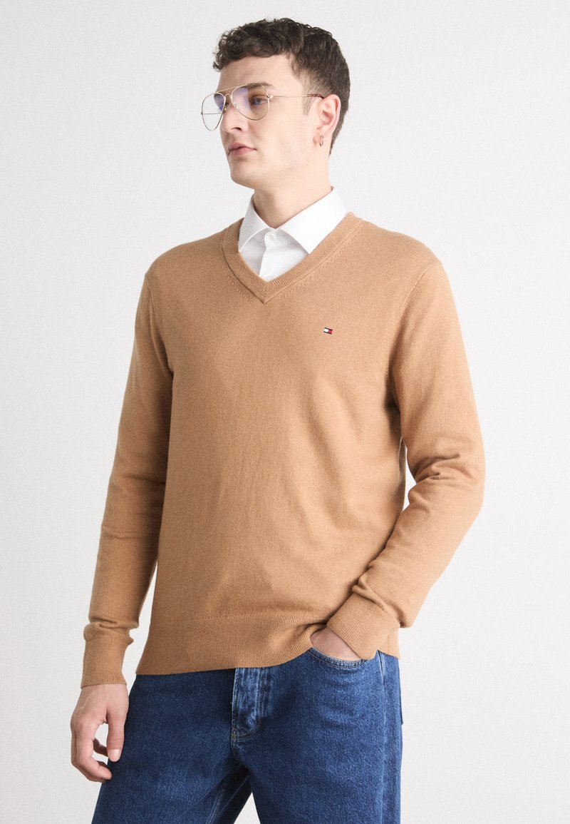 Maglione color cammello a V, realizzato in un tessuto morbido. Presenta maniche lunghe e polsini a costine. Piccolo logo sul lato sinistro del petto. Indossato sopra una camicia bianca.