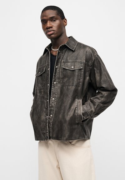 Άνδρας που φορά σκούρο πλυντήριο με κουμπιά πάνω από μαύρο πουκάμισο και ανοιχτό μπεζ παντελόνι, στέκεται με τα χέρια στις τσέπες απέναντι σε απλό φόντο.