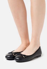 Czarne baletki wykonane z gładkiej skóry, z okrągłym noskiem, smukłym detalem kokardy oraz małymi złotymi akcentami na końcach kokardy.