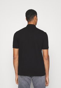 Homme portant un polo noir uni à manches courtes et un jean gris, se tenant de dos contre un fond blanc uni.