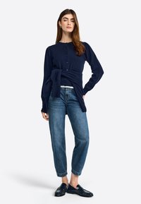 Marineblaue Strickjacke mit rundem Ausschnitt, knöpfigem Vorderteil und gebundenem Bund, kombiniert mit lässig geschnittenen Jeans und dunklen Mokassins.