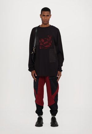 Schwarzes Langarmhemd mit burgunderfarbenen Musterakzenten und einer Tasche; kombiniert mit roten und schwarzen, taillierten Hosen mit eckigen Designdetails.