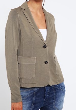 Femme portant un blazer vert olive avec deux boutons et de grandes poches avant à rabat, associé à un jean bleu et un haut blanc.