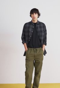 Black and gray plaid button-up shirt over a black t-shirt, paired with olive green drawstring pants. Simple design and relaxed fit.