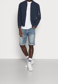 Man som bär en marinblå bomberjacka, en vit tryckt t-shirt, uppvikta denimshorts, vita strumpor med svarta ränder och vita sneakers, står med handen i fickan.