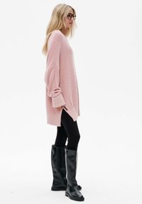 Lichtroze oversized sweater met lange mouwen, geribbelde manchetten en zijslitsen, gecombineerd met zwarte leggings en kniehoge zwarte laarzen.
