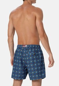 Uomo che indossa pantaloni da bagno blu a fantasia con vita elastica e tasca posteriore, in piedi senza maglietta di spalle su uno sfondo neutro.