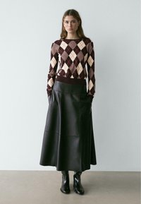 Braun-cremefarbenes Rautenmuster-Pullover mit eng anliegenden Bündchen, kombiniert mit einem langen schwarzen Lederminirock und schwarzen Stiefeletten. Glatte Texturen und geschichtetes Design.