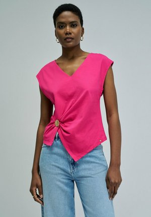 Mujer con top sin mangas de color rosa brillante con detalle de botones delanteros y vaqueros azul claro, de pie frente a un fondo gris liso.