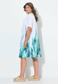 Kurzärmliges weißes Kleid mit einem türkisfarbenen Pflanzenmuster am Saum, in lockerer Passform. Getragen mit hellbraunen flachen Sandalen.