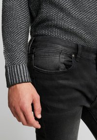 Musta denimpaita on kapealahkeinen ja siinä on viiden taskun malli. Yhdistetty harmaaseen, teksturoituun neulepaitaan, jossa on resorireunus.