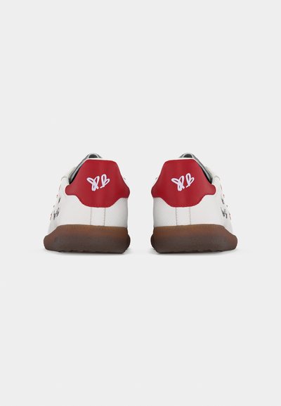 Λευκά αθλητικά παπούτσια με καφέ σόλες και κόκκινες ταμπελίτσες στη φτέρνα που φέρουν λευκό λογότυπο πεταλούδας, εμφανιζόμενα από πί σω σε λευκό φόντο.