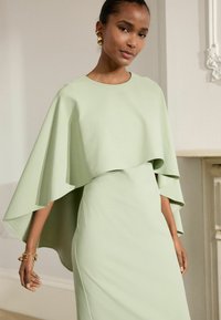 Robe vert clair avec un design de type cape. Texture lisse, silhouette ajustée et manches en couches. Un bracelet doré accessoirise le look.