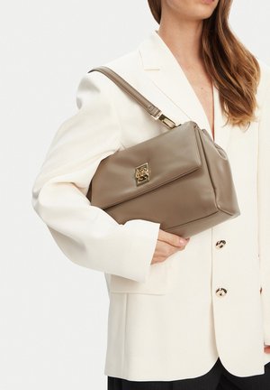 Žena v krémovém sáčku držící tašku přes rameno z taupe kůže se zlatou sponou na světlém pozadí.