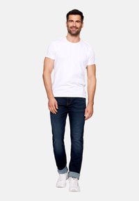 Camiseta blanca de manga corta combinada con jeans azul oscuro y zapatillas blancas; el modelo está de pie de forma casual con las manos relajadas a los lados.