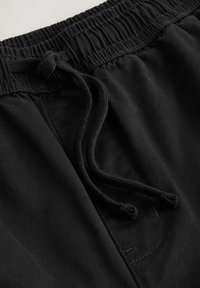 Pantalon en tissu noir avec taille élastique et cordon de serrage noué, montrant des coutures détaillées et la texture.