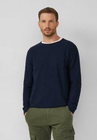 Marineblauer Strickpullover mit strukturiertem Finish und weißem Kragenbesatz, kombiniert mit olivgrünen Cargo-Hosen. Das Kleidungsstück hat lange Ärmel und gerippte Bündchen.