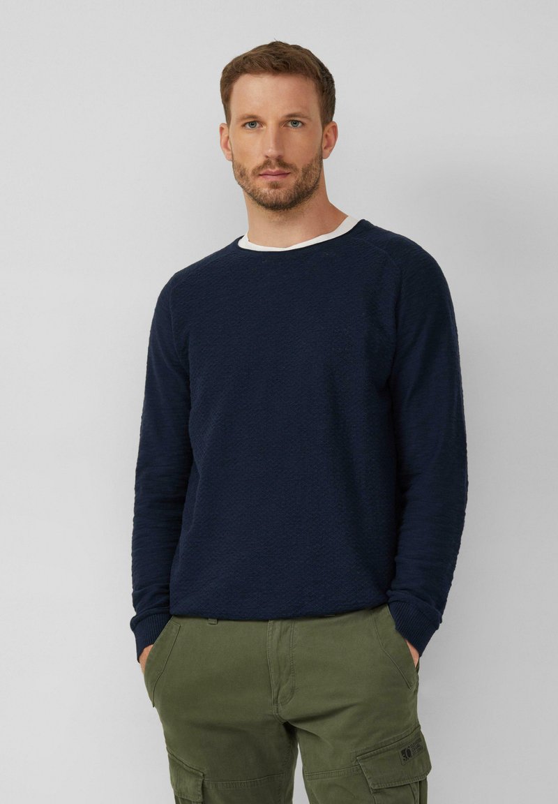 Marineblauer Strickpullover mit strukturiertem Finish und weißem Kragenbesatz, kombiniert mit olivgrünen Cargo-Hosen. Das Kleidungsstück hat lange Ärmel und gerippte Bündchen.