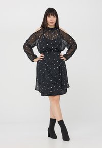 Vestito nero a pois con maniche lunghe trasparenti, colletto alto e spalle scavate. Si adatta sopra il ginocchio. Abbinato a stivali neri alla caviglia.