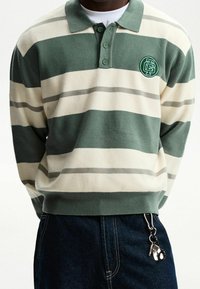 Person klädd i en grön och vit randig långärmad polo-tröja med mörka jeans och en nyckelring fäst vid midjan.