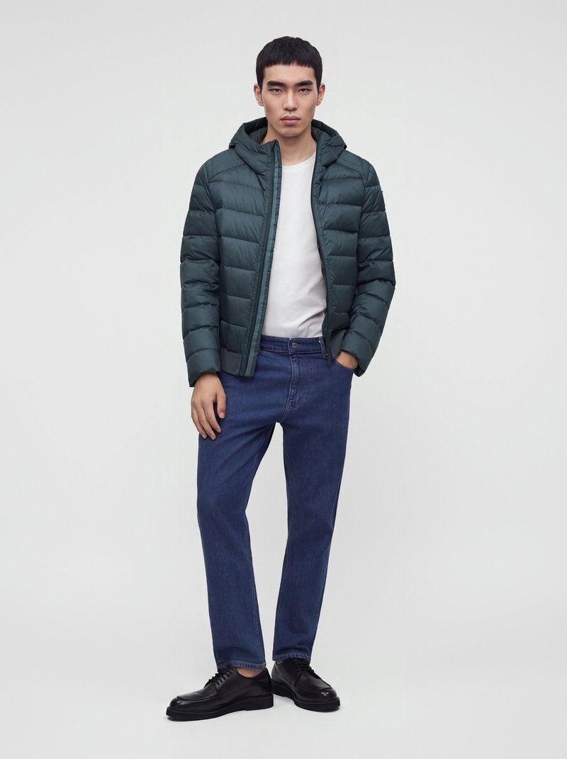 Jeune homme portant une doudoune vert foncé sur une chemise blanche, un jean bleu et des chaussures noires, debout devant un fond clair uni.