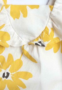 Tela blanca con un gran patrón floral amarillo. Presenta un cuello con volantes y ligeras frunces en la parte del cuello. Material de algodón texturizado.