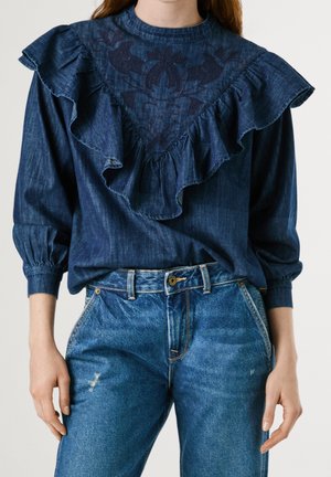 Kvinde iført en mørkeblå bluse med flæser og broderi samt lyseblå højhælede jeans med let slid.