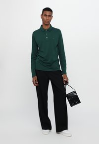 Ung mann i mørkegrønn langermet polo, svarte vide bukser, hvite sneakers, holder en liten svart Marni-veske i reim.