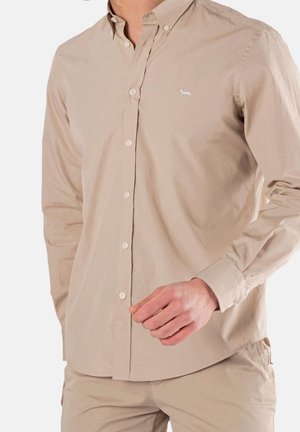 Mężczyzna noszący beżową koszulę na długi rękaw z małym białym logo psa na piersi i dopasowane beżowe spodnie, ręka blisko talii.