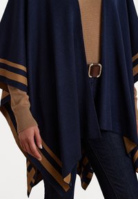 Poncho lavorato a maglia a righe blu navy e marrone sopra una camicia a maniche lunghe beige, jeans scuri e cintura marrone con fibbia in argento, mano rilassata al lato.