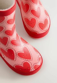 Rosa Gummistiefel mit roten Herzmustern und gezackten Kanten, mit einer glatten Oberfläche und strukturierten roten Sohlen.