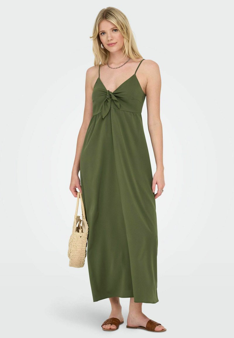 Robe maxi en tissu vert olive avec un design noué au niveau de la poitrine, des fines bretelles et une coupe ample. Associée à un sac en osier et des sandales.