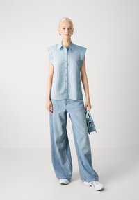 Camicetta senza maniche di colore blu chiaro con righe verticali, chiusura davanti con bottoni, abbinata a jeans a gamba larga blu chiaro e una piccola borsa coordinata.