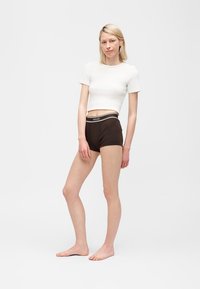 Νεαρή γυναίκα στέκεται ξυπόλητη φορώντας ένα λευκό κοντό t-shirt και μαύρα σορτς Versace μπροστά σε έναν απλό ανοιχτόχρωμο φόντο.