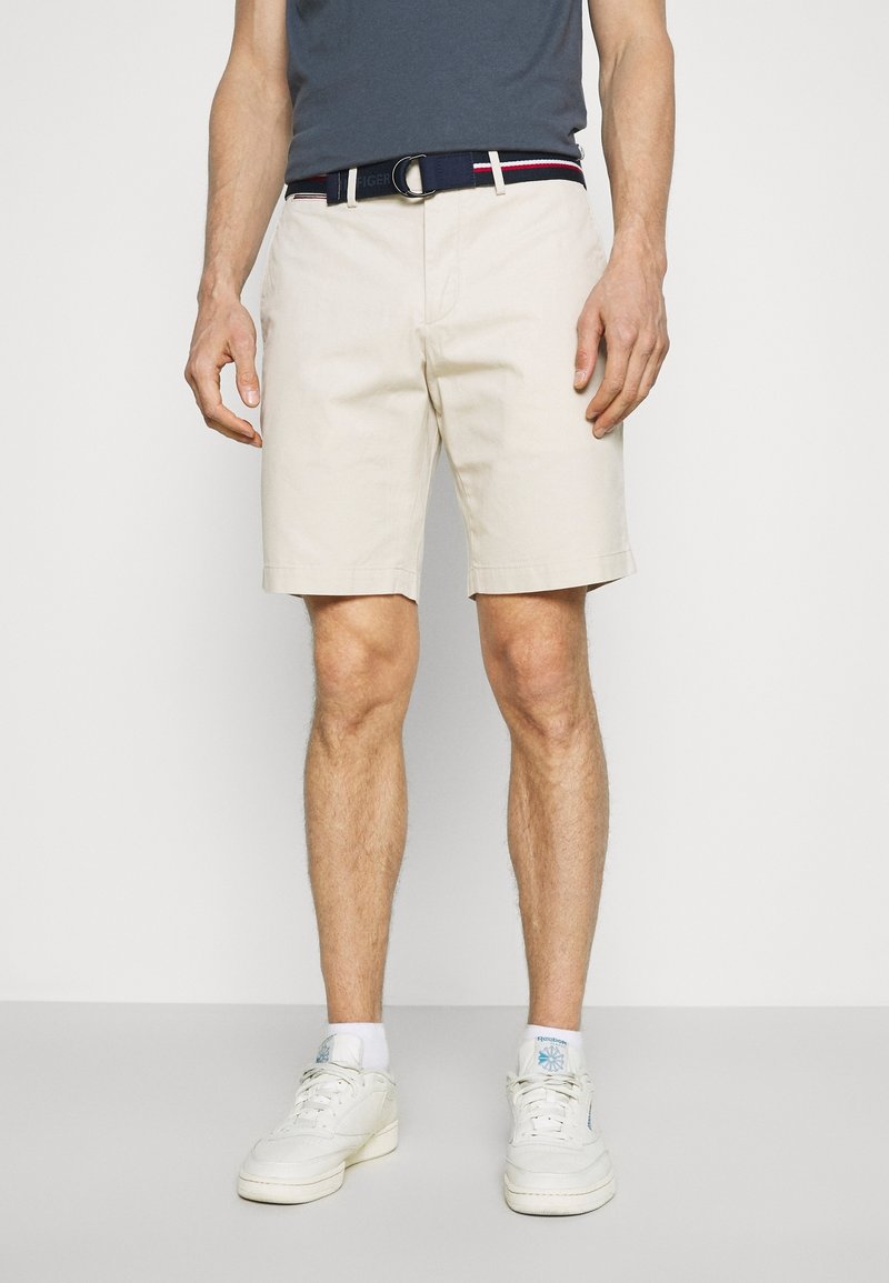 Beige puuvillahousut suoran leikkauksen kanssa, käytössä tummansininen vyö, jossa on punaisia ja valkoisia yksityiskohtia, yhdistetty valkoisiin tennareihin.