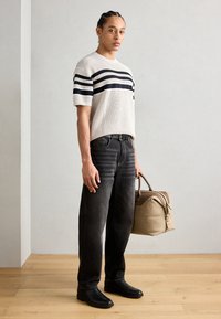 Helle beige Strickjacke mit marineblauen Streifen, schwarze Jeans und schwarze Schuhe, hält eine sandfarbene Reisetasche mit braunen Akzenten.