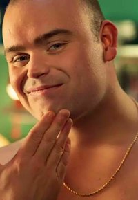 Un uomo con i capelli corti si tocca il mento, indossando una sottile collana d'oro attorcigliata. Lo sfondo presenta un'ambientazione sfocata e colorata.