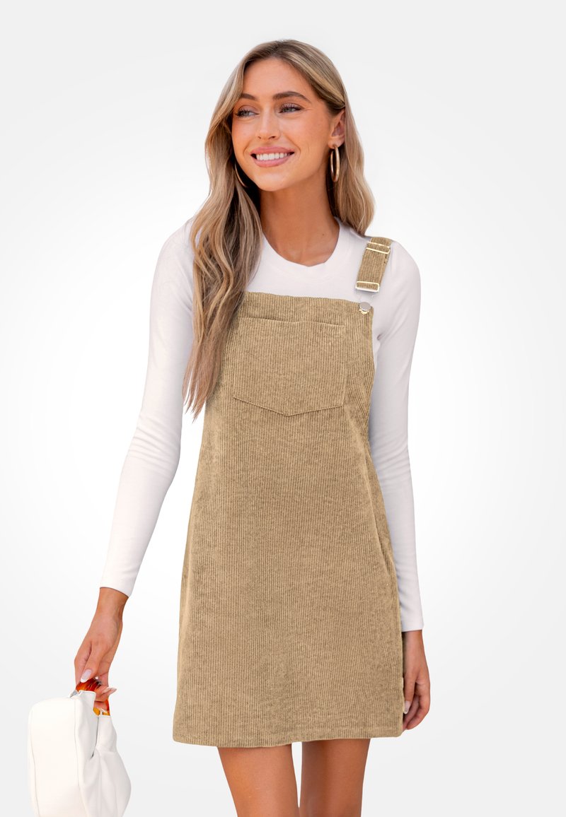 Beige Corduroy-Latzkleid mit großer Fronttasche, verstellbaren Trägern und gerillter Textur. Getragen über einem weißen Langarmshirt.