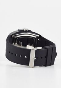 Bracelet de montre en silicone noir avec plusieurs trous, boucle en métal argenté, et un boîtier carré avec une finition mate, mettant en valeur un fond en verre.