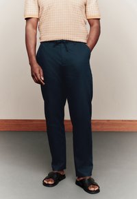 Lühikeste varrukatega mustriline särk soojades toonides, kombineeritud tumedate mereväe pükstega, millel on taskud, ja mustade laia rihmaga tasaste sandaalidega.