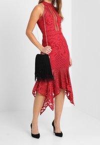 Frau in einem ärmellosen roten Spitzenkleid mit asymmetrischem Saum, die eine schwarze Fransenhandtasche hält und schwarze High Heels trägt.