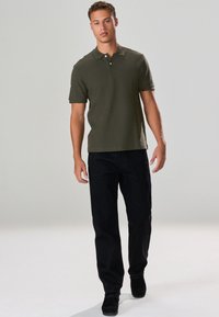 Maslinasto zelena polo majica s ovratnikom i dva gumba, uparena s pripijenim crnim trapericama. Topli ton kože i casual crne cipele.