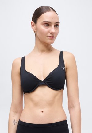 Tânără cu părul dat pe spate purtând un top de bikini negru cu un mic logo alb pe partea stângă, stând în fața unui fundal simplu.