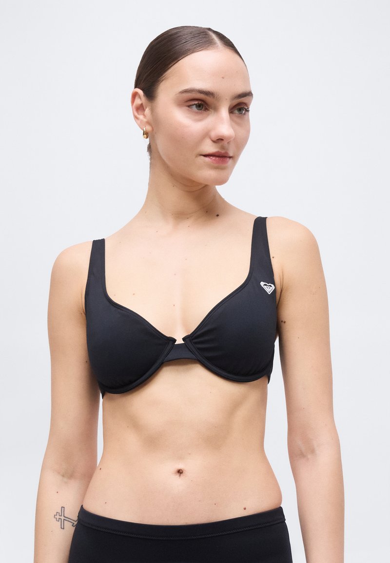 Jeune femme aux cheveux lissés en arrière portant un haut de bikini noir avec un petit logo blanc sur le côté gauche, debout devant un fond uni.