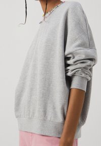 Sudadera gris con cuello redondo, corte holgado y puños fruncidos. El cuello cuenta con acentos de cuentas coloridas. Tejido suave y texturizado.