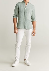 Man in een opgerolde lichtgroene blouse met knopen, witte broek en witte sneakers tegen een effen beige achtergrond.