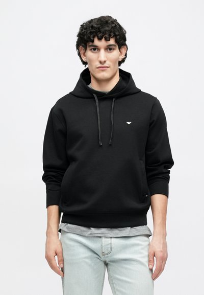 Melnā kapuce no mīksta auduma ar priekšējo kabatu, aukliņu kapucē un nelielu logotipu krūtīs. Valsts kopā ar gaiši zilām džinsiem.