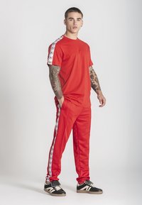 Mladý muž s tetovaniami, oblečený v červenom športovom obleku s bielymi pruhmi s logom na rukávoch a nohaviciach, stojaci s jednou rukou v vrecku.