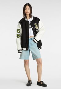Sort og hvid varsity-jakke med grøn broderi, trykknapper og striber; parret med lyseblå denimshorts. Sorte slip-on sko.