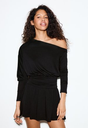 Kobieta o kręconych włosach, ubrana w czarną, długą sukienkę z odkrytymi ramionami, stojąca na białym tle.